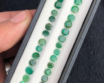 May include: A selection of round, faceted emerald gemstones in various shades of green. The gems are arranged in two parallel rows within a black display box with a white interior. The gemstones are of different sizes.