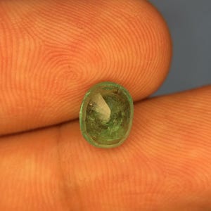 May include: A small, oval-shaped, translucent green gemstone with a faceted surface. The stone displays natural internal inclusions and variations in colour intensity, ranging from light to deep forest green throughout its crystalline structure.