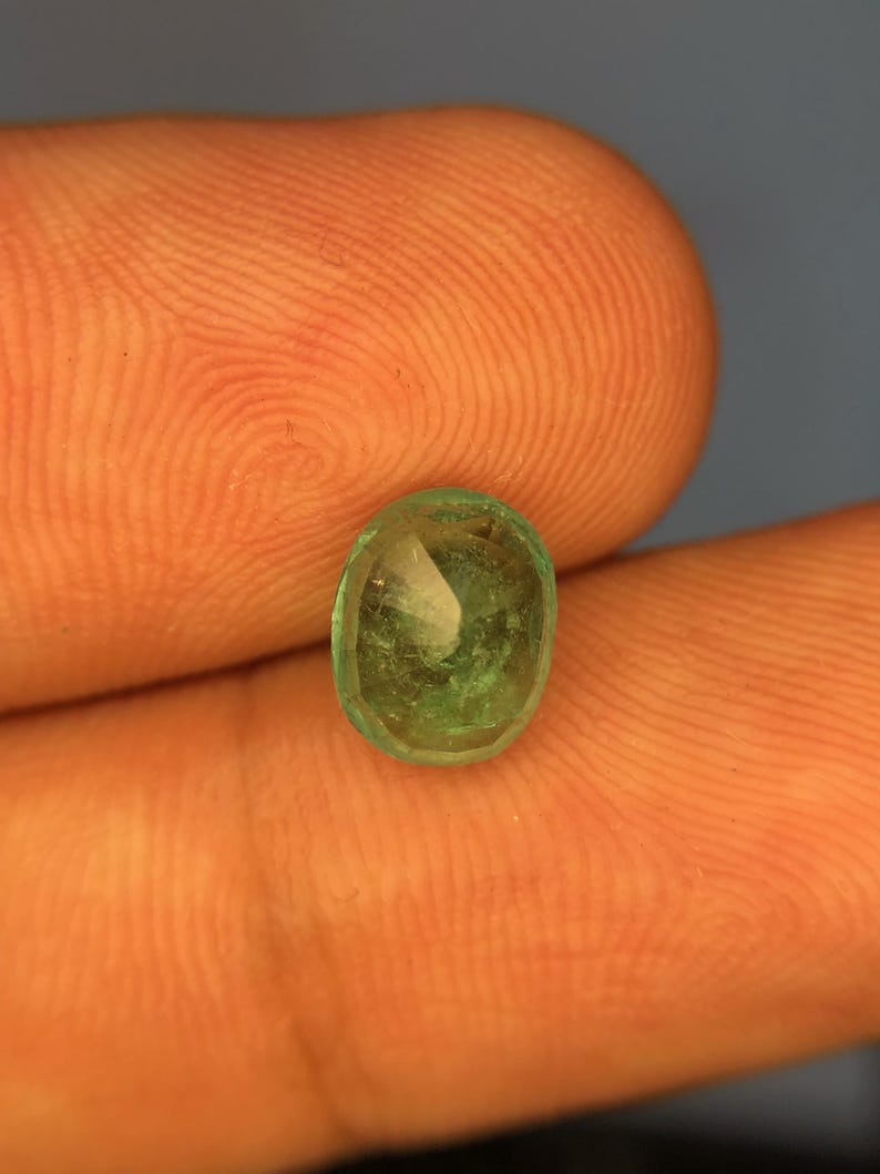 May include: A small, oval-shaped, translucent green gemstone with a faceted surface. The stone displays natural internal inclusions and variations in colour intensity, ranging from light to deep forest green throughout its crystalline structure.