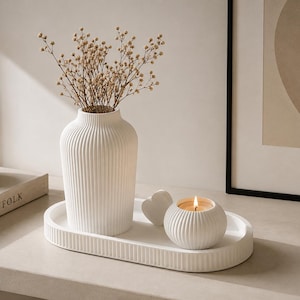 Peut inclure: Un vase en céramique blanche cannelée, un bougeoir cannelé assorti avec une bougie allumée et un petit objet en forme de cœur disposés sur un plateau ovale cannelé. Le vase contient un bouquet de fleurs séchées beiges, formant un ensemble.