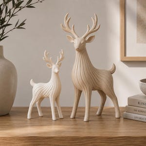 Pode incluir: Duas estatuetas de veado na cor creme com texturas estriadas verticais suaves nos corpos. O veado maior está à direita com chifres altos e ramificados, enquanto o veado menor está à esquerda com chifres mais curtos, ambos voltados para a frente.