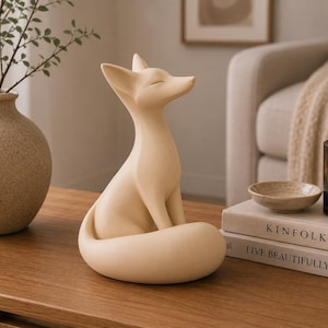 May include: A smooth, matte beige ceramic fox figurine in a seated position with its tail wrapped around its base. The fox has pointed ears, closed eyes, and a slender snout, presented as a solid, monochromatic decorative sculpture.