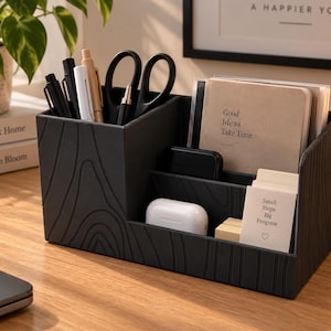Könnte beinhalten: Ein schwarzer Schreibtisch-Organizer mit texturiertem Holzmaserungsmuster und mehreren Fächern. Er enthält Stifte, eine Schere, ein weißes Ladegehäuse und Notizbücher mit dem Text Good Ideas Take Time und Small Steps Big Progress mit Herzsymbol.
