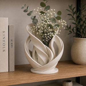 Può includere: Un vaso in ceramica bianco sporco opaco con un design scultoreo e vorticoso caratterizzato da pannelli curvi stratificati che formano una struttura circolare aperta. Il vaso contiene un bouquet di foglie di eucalipto e piccoli fiori bianchi.