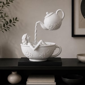 May include: A white matte sculpture featuring a person reclining inside a large teacup while a teapot suspended above pours liquid into the cup. The teacup and teapot are decorated with embossed floral patterns on their exterior surfaces.