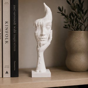 Puede incluir: Una escultura blanca de un rostro humano que descansa sobre una mano, con un efecto de derretimiento o goteo en un lado de la cara. La figura está montada sobre una base cuadrada y se coloca en posición vertical, mostrando rasgos suaves y calmados.