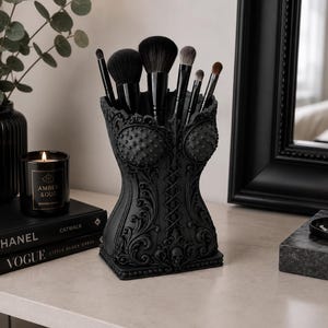 Könnte beinhalten: Ein mattschwarzer Make-up-Pinselhalter in Korsettform mit filigranen Schnörkeln, einer zentralen Schnürung und einem kleinen Totenkopf-Detail am Sockel. Der Behälter enthält sechs Make-up-Pinsel mit schwarzen Griffen in verschiedenen Größen.