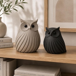 May include: Two owl figurines with a smooth, ribbed texture and round black eyes. One owl is light beige and the other is matte black. Both figures feature pointed ear tufts and a rounded body shape, displayed as a pair on a wooden surface.