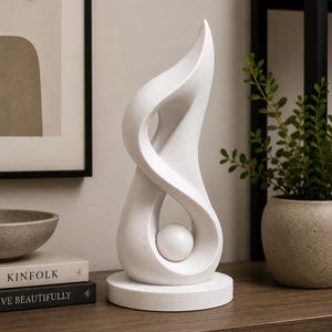Könnte beinhalten: Eine weiße abstrakte Skulptur mit einer glatten, flammenartigen vertikalen Form, die sich von einem runden Sockel nach oben wölbt, mit einer kleinen, massiven weißen Kugel, die in der zentralen Öffnung der texturierten, matten Struktur schwebt.
