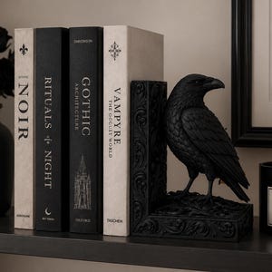 Puede incluir: Un sujetalibros de cuervo negro con detalles tallados intrincados, colocado junto a cuatro libros titulados Noir, Rituals of Night, Gothic Architecture y Vampyre. El cuervo se apoya sobre una base rectangular con relieves florales.