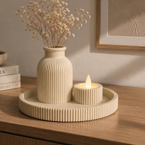 Puede incluir: Una bandeja circular de color beige con borde acanalado sostiene un jarrón acanalado a juego con flores secas de paniculata y un pequeño portavelas cilíndrico acanalado con una vela LED encendida en el centro.
