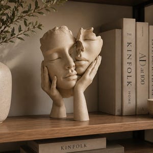 May include: A beige sculpture depicting two faces joined together, with one face resting in a pair of hands. The sculpture has a matte, textured finish and is positioned between a speckled vase and several books with visible text on their spines.