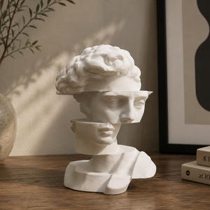 Può includere: Una scultura bianca di una testa e spalle umane divise in tre sezioni orizzontali sfalsate. Il pezzo presenta lineamenti classici e capelli strutturati, con una finitura opaca liscia, creando un aspetto frammentato del busto.