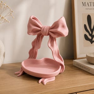 Peut inclure: Un plateau décoratif rose mat doté d'un bassin central en forme de cœur soutenu par une structure en ruban sculpté. Le design comprend un grand nœud proéminent sur le dessus avec des extrémités de ruban fluides formant la base du plateau.