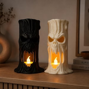 May include: Two cylindrical tree trunk sculptures, one matte black and one off-white, each featuring a carved, screaming face with hollow eyes and an open mouth. Inside each hollow mouth sits a small, glowing, warm-toned LED tea light candle.