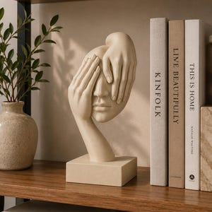 May include: A beige sculpture of a human face partially covered by two hands, mounted on a rectangular base. The sculpture features smooth, matte surfaces and detailed fingers resting over the eyes and forehead, creating a contemplative expression.