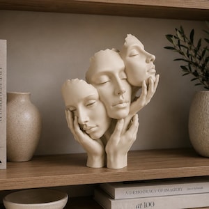 Könnte beinhalten: Eine cremefarbene Skulptur, die drei menschliche Gesichter zeigt, die mit Händen verschlungen sind. Das zentrale Gesicht wird von zwei weiteren flankiert, wobei Hände um die Kinne und Wangen der Gesichter positioniert sind. Die Skulptur hat eine glatte, matte Oberfläche.