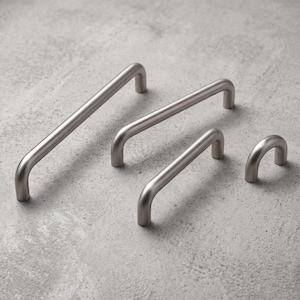 Könnte beinhalten: Ein Set aus vier Schrankgriffen aus gebürstetem silberfarbenem Metall und einem passenden Knopf, die alle ein glattes, abgerundetes Bogendesign aufweisen. Die Beschläge variieren in der Länge, von einem langen Griff bis zu einem kleinen Knopf.