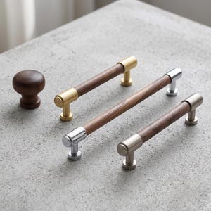 May include: A collection of cabinet hardware including one round dark wood knob and three cylindrical drawer pulls. Each pull features a central dark wood bar with metallic end caps in brushed gold, polished chrome, and brushed nickel finishes.