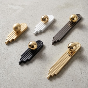 Può includere: Cinque maniglie per armadietti allungate con una base a gradini e un pomello sferico liscio sulla parte superiore. Il set include varianti con finiture in oro opaco, bianco, marrone scuro, nero opaco e oro lucido.