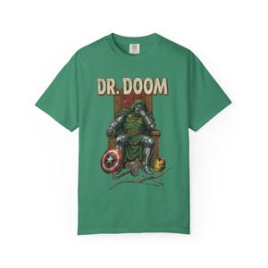 May include: Green t-shirt featuring a comic book illustration of Dr. Doom seated on a throne. The text "DR. DOOM" is above the illustration. The image includes Captain America's shield, Thor's hammer, and Iron Man's helmet.