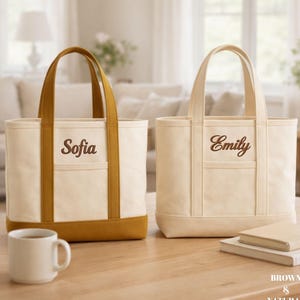 May include: Two canvas tote bags with the names "Sofia" and "Emily" embroidered on the front. The bags are cream-colored with brown accents and handles. A white mug and a stack of books sit nearby.