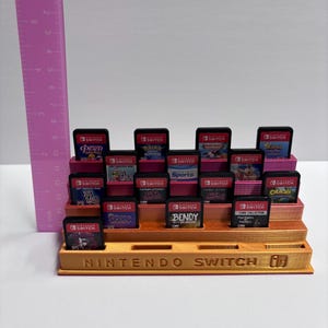 May include: A 3D-printed, tiered display stand for Nintendo Switch game cartridges. The stand is a light brown color and features the words "NINTENDO SWITCH" on the front. The stand holds multiple game cartridges, each with a different game title. The ruler shows the stand is approximately 6 inches tall.
