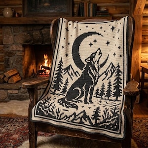 Pode incluir: Uma manta de crochê na cor creme com um design gráfico preto de um lobo uivando para uma lua crescente, cercado por estrelas, pinheiros e silhuetas de montanhas, sobre uma poltrona de madeira em uma sala com lareira de pedra.