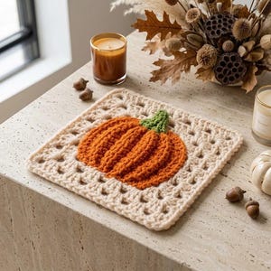 May include: A square crochet granny square coaster featuring a textured orange pumpkin with a green stem centred on a cream-coloured yarn base. The pumpkin has raised vertical ridges and is crafted with a classic open-work stitch pattern.