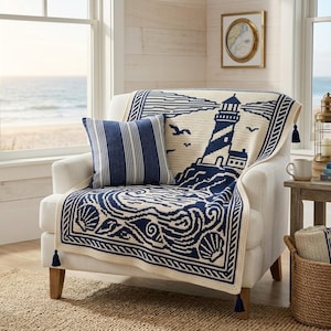 Könnte beinhalten: Ein cremefarbener Sessel, der mit einer marineblau-cremefarbenen Strickdecke mit Leuchtturm-, Meereswellen- und Muschelmotiven bedeckt ist. Ein marineblau-weiß gestreiftes Kissen liegt auf der Sitzfläche und ist mit marineblauen Quasten verziert.