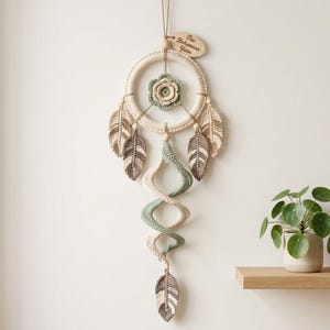 May include: A wall hanging featuring a circular frame wrapped in cream yarn with a central sage green crocheted flower. Four crocheted leaves in cream and brown hang from the sides, and a long, spiraling cream and sage green tail ends in a leaf.