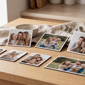 May include: A set of square photo coasters displaying family portraits. The coasters are arranged on a wooden table, showing images of a smiling family. The photos have white borders and dark edges.