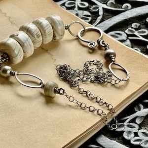 May include: A necklace featuring large, off-white, cylindrical beads with dark lines, connected by silver-toned metal rings and a chain. The necklace rests on a vintage paper surface, with a dark, carved wooden background.
