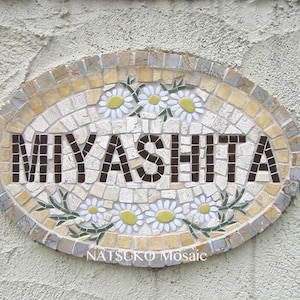 Op de afbeelding: Ovaal mozaïeken bord met de naam MIYASHITA in donkerbruine blokletters. Het bord is versierd met witte madeliefjes met gele centra en groene bladeren. De tekst "NATSUKO Mosaic" staat onderaan.