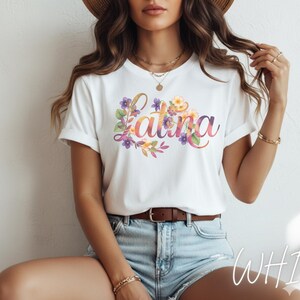 May include: White t-shirt with the word "Latina" in a colorful, floral design. The text is in a script font, surrounded by watercolor flowers in shades of purple, yellow, and orange. The shirt is paired with denim shorts.