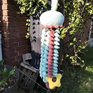 May include: A handmade crochet jellyfish mobile in pastel colors. The jellyfish has a light blue head, a pink and blue body, and a yellow star-shaped teether at the bottom. The mobile is hanging outdoors near a brick wall and greenery.