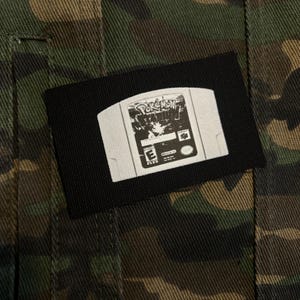May include: A black patch featuring a white image of a Nintendo 64 video game cartridge. The cartridge has the Pokemon logo and other text. The patch is on a camouflage jacket.
