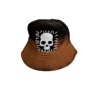 Pode incluir: Um chapéu de balde castanho e preto com um desenho de caveira branca. O chapéu tem um efeito gradiente, com a parte superior preta desvanecendo-se em castanho. A caveira é branca com um contorno branco e está centrada na frente.