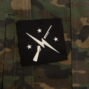 Könnte beinhalten: Tarnjacke mit einem schwarzen quadratischen Aufnäher. Der Aufnäher zeigt eine weiße Grafik einer Pistole, eines Blitzes und Sterne. Der Stoff der Jacke weist Farbtöne von Grün, Braun und Schwarz auf.