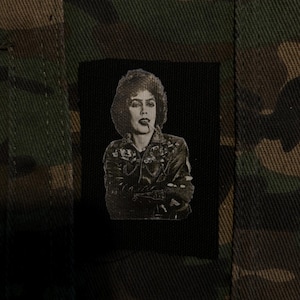 Op de afbeelding: Een rechthoekige zwarte stoffen patch met een wit zeefdrukportret van een persoon met donker krullend haar, donkere lippenstift en een leren jas, gecentreerd op een textielachtergrond met een groen, bruin en zwart camouflagepatroon.