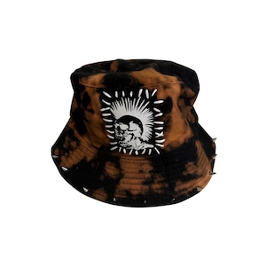 Pode incluir: Um chapéu bucket tie-dye preto e castanho com pontas prateadas à volta da aba. O chapéu apresenta um patch branco com um gráfico a preto e branco de uma caveira com um moicano.