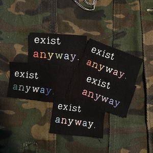 May include: Black patches with the words "exist anyway" embroidered in colorful thread. The patches are displayed on a camouflage jacket. A silver chain and safety pin are visible at the top.