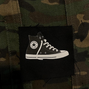 May include: A square black fabric patch featuring a white graphic of a high-top sneaker with the Converse All Star logo, stitched onto a green, brown, and black camouflage patterned textile background with visible vertical seams.