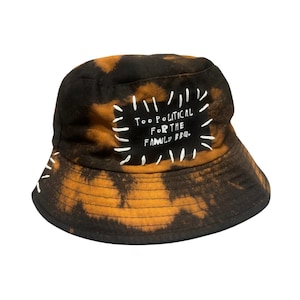 Puede incluir: Un sombrero de pescador tie-dye negro y naranja con un parche rectangular de bordes cosidos a mano en blanco. El parche muestra el texto "TOO POLITICAL FOR THE FAMILY BBQ." en letras blancas, con detalles de costuras blancas en el ala.