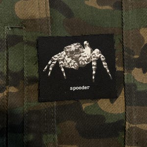 Könnte beinhalten: Ein schwarzer Aufnäher mit einer detaillierten weißen Spinnenillustration und dem Wort "spooder". Der Aufnäher ist auf einem Camouflage-Stoff in Grün-, Braun- und Schwarztönen aufgenäht.