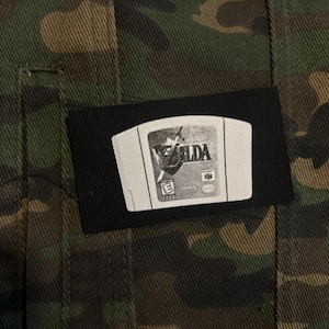 Puede incluir: Un parche negro con una imagen blanca de un cartucho de videojuego de Nintendo 64 con la palabra "ZELDA" impresa. El parche está cosido a una chaqueta de camuflaje.