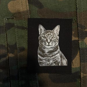 Peut inclure: Un écusson noir avec une illustration en niveaux de gris d'un chat avec un troisième œil, cousu sur un tissu camouflage. Le chat est en position assise, regardant vers l'avant. Le tissu présente des tons verts, marron et noirs.
