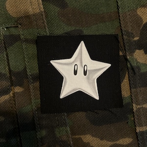 May include: A close-up of a camouflage jacket with a black square patch. The patch features a white star with two black oval eyes, reminiscent of a character from a video game.