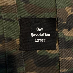 May include: Camouflage-patterned jacket with a black patch. The patch features the words "One Revolution Later" in white lettering. The jacket's fabric has a mix of green, brown, and tan colors.