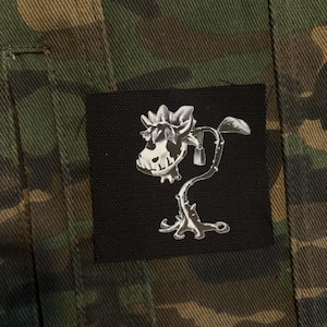 Puede incluir: Chaqueta de camuflaje con un parche cuadrado negro. El parche presenta una ilustración blanca de una flor con forma de calavera con espinas y hojas. La tela de la chaqueta tiene un patrón verde, marrón y beige.
